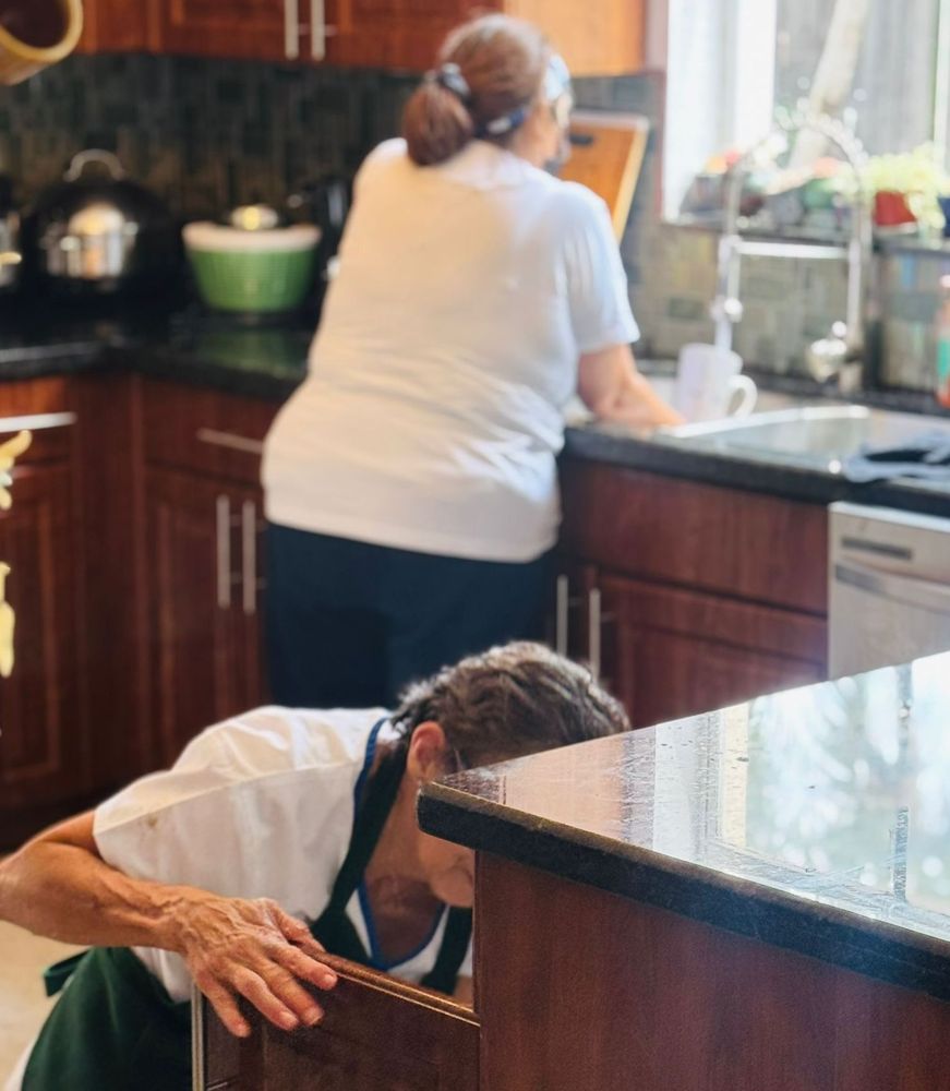 Two woman cleaning a kitchen; one doing dishes and the other tending to a cabinet
