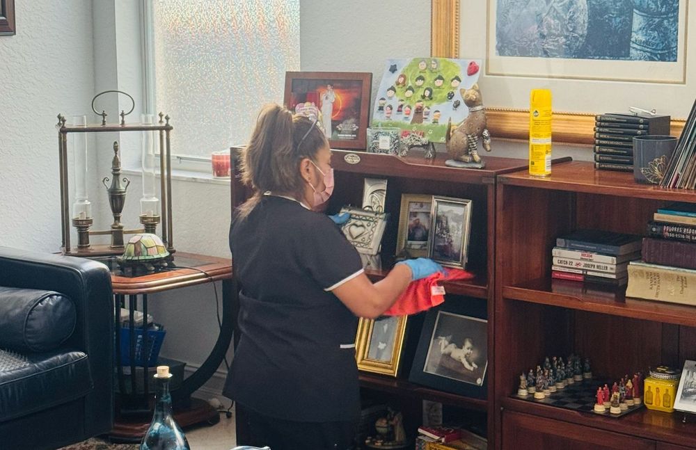 A woman in a black cleaning outfit kneeling in front of a shelf, meticulously dusting picture frames