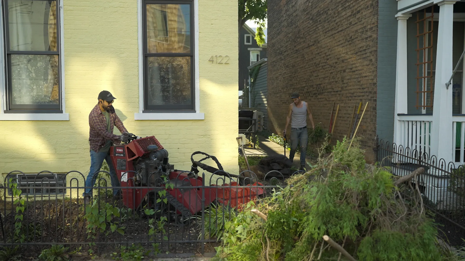 Two landscapers in front of a yellow house; one pushing a red machine and the other carrying a small potted plant